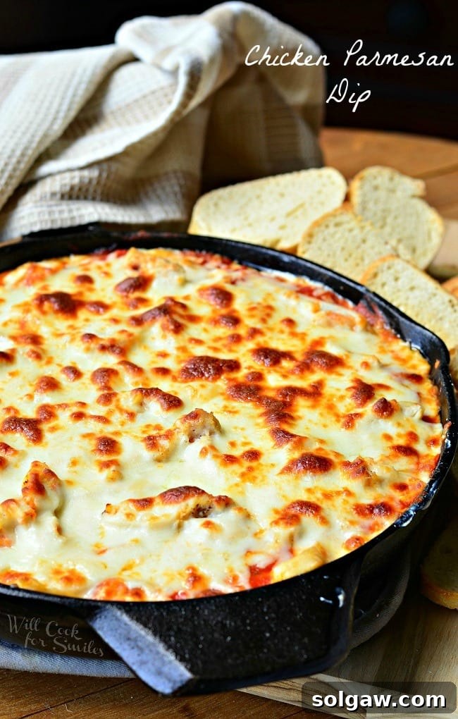 A whole black cast iron skillet filled with golden brown, bubbly Chicken Parmesan Dip, with a scattered arrangement of toasted baguette slices in the background.