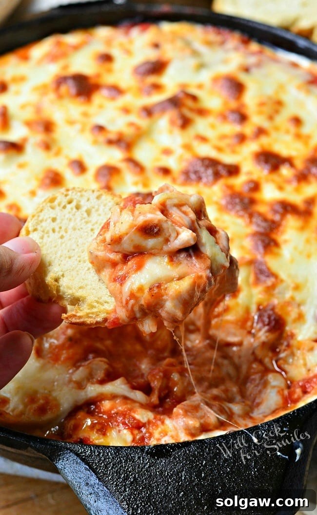 A hand holding a piece of crispy baguette, dipping it into the warm and stringy Chicken Parmesan Dip in a black skillet, showing off the gooey cheese pull.