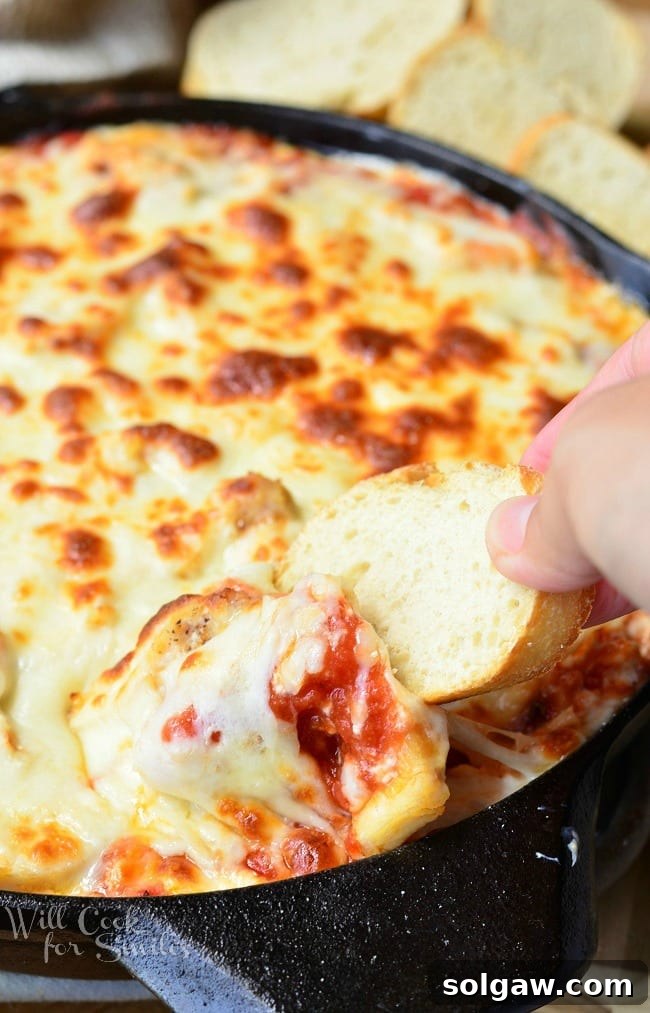 Another hand reaching into a black skillet filled with bubbling Chicken Parmesan Dip, scooping up a portion with a piece of toasted bread.