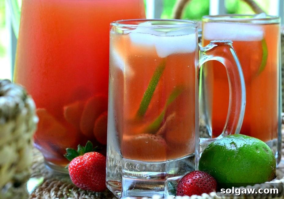 Close-up side view of a glass mug filled with Strawberry Lime Infused Iced Tea, showing the vibrant colors of strawberry and lime slices amidst the ice. Another mug and a pitcher are in the background, subtly out of focus, enhancing the inviting atmosphere.