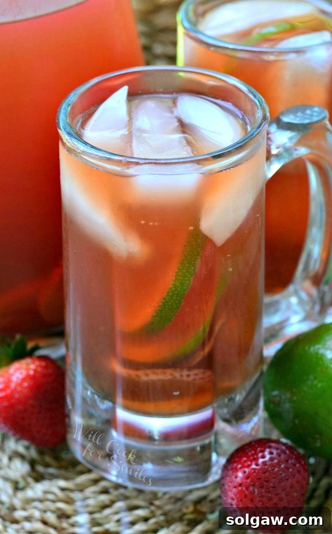 Clear glass mug filled with strawberry lime infused iced tea, garnished with fresh fruit, set in front of a pitcher and another mug of tea.