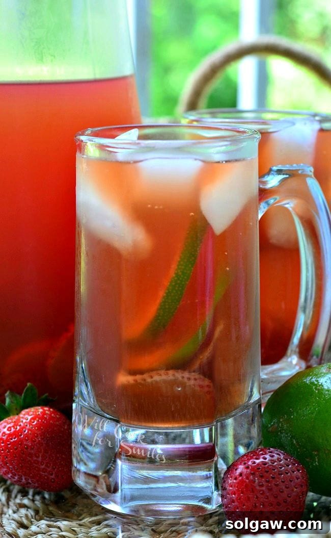 Overhead view of a glass mug of Strawberry Lime Infused Iced Tea, showing ice, strawberry, and lime slices. A pitcher and another mug are in the background, along with loose strawberries and lime on the table, creating an inviting summer scene.