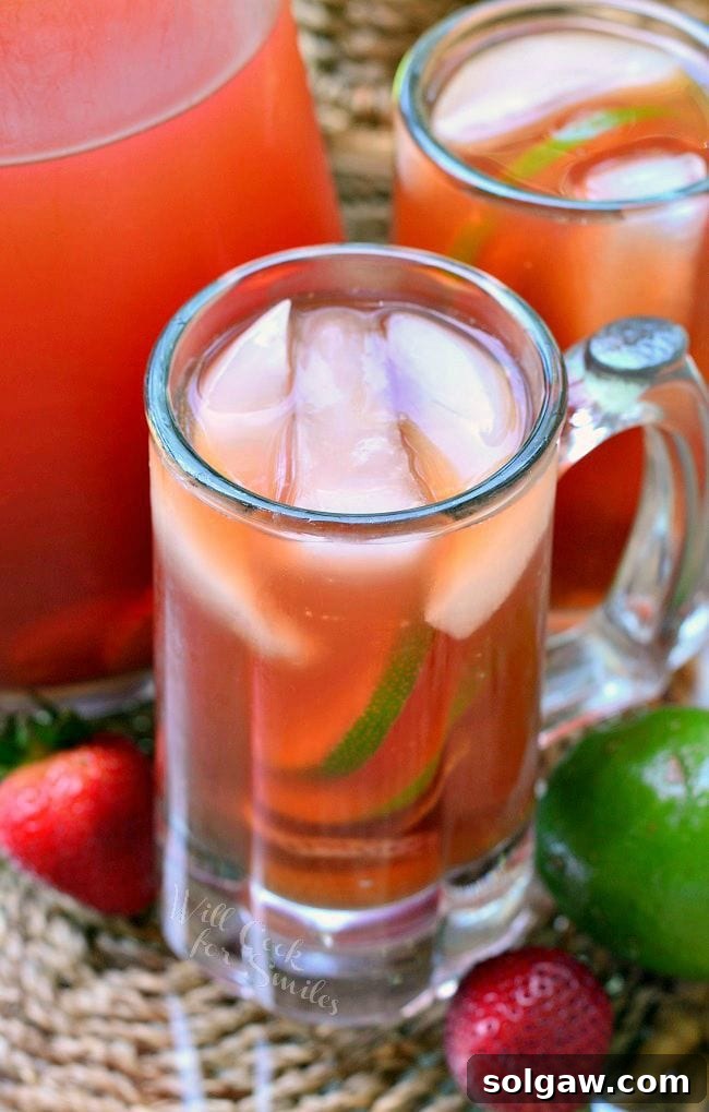 Top-down view of a glass of Strawberry Lime Infused Iced Tea, showing the arrangement of ice, strawberry, and lime slices. The background features another mug and a pitcher, along with fresh fruit, creating an appealing and refreshing visual.