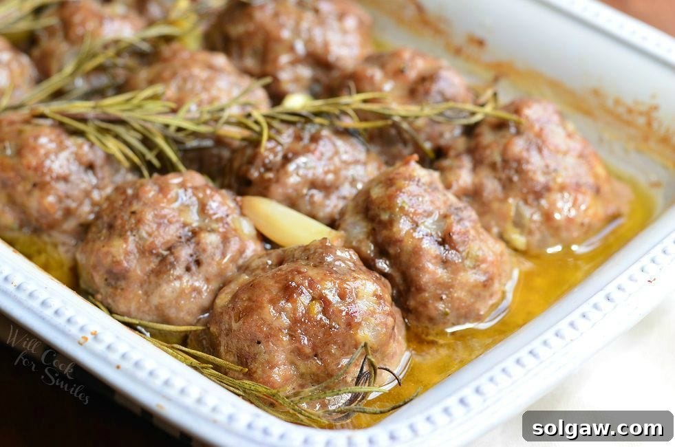 close up view of roasted garlic rosemary baked meatballs in a white baking dish on a wooden table topped with 3 rosemary sprigs