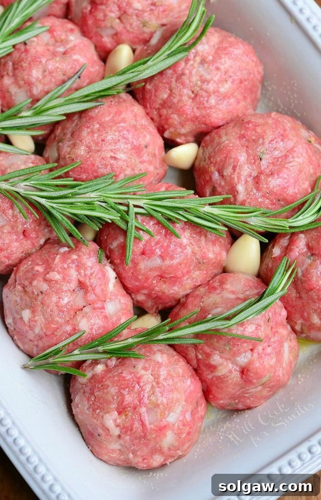 Uncooked meatballs in a white baking dish with rosemary and pieces of garlic.