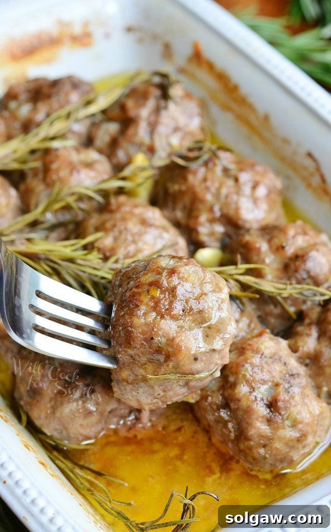 Rosemary Garlic Baked Meatballs in a white baking dish with rosemary and pieces of garlic after being cooked. A fork is inserted into one meatball holding it above the others.