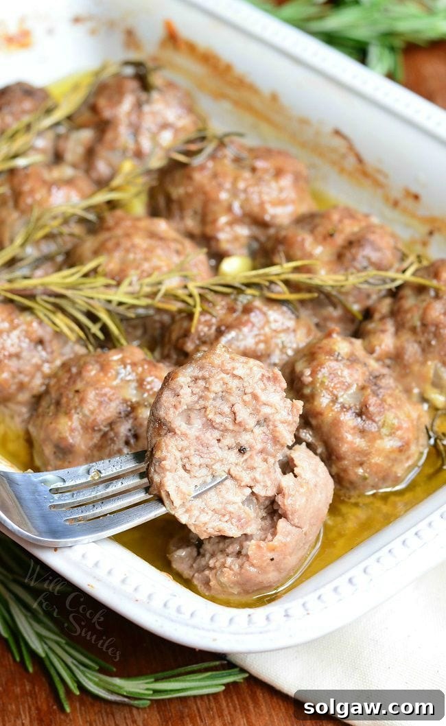 Rosemary Garlic Baked Meatballs in a white baking dish with rosemary and pieces of garlic after being cooked. A fork is holding up half a meatball above the dish.