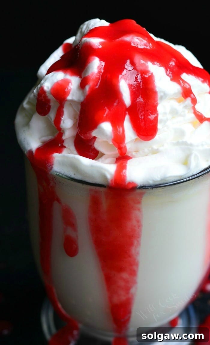 Clear glass coffee mug filled with white chocolate raspberry halloween hot chocolate on a black table with raspberry sauce splatter on table around base of glass viewed close up