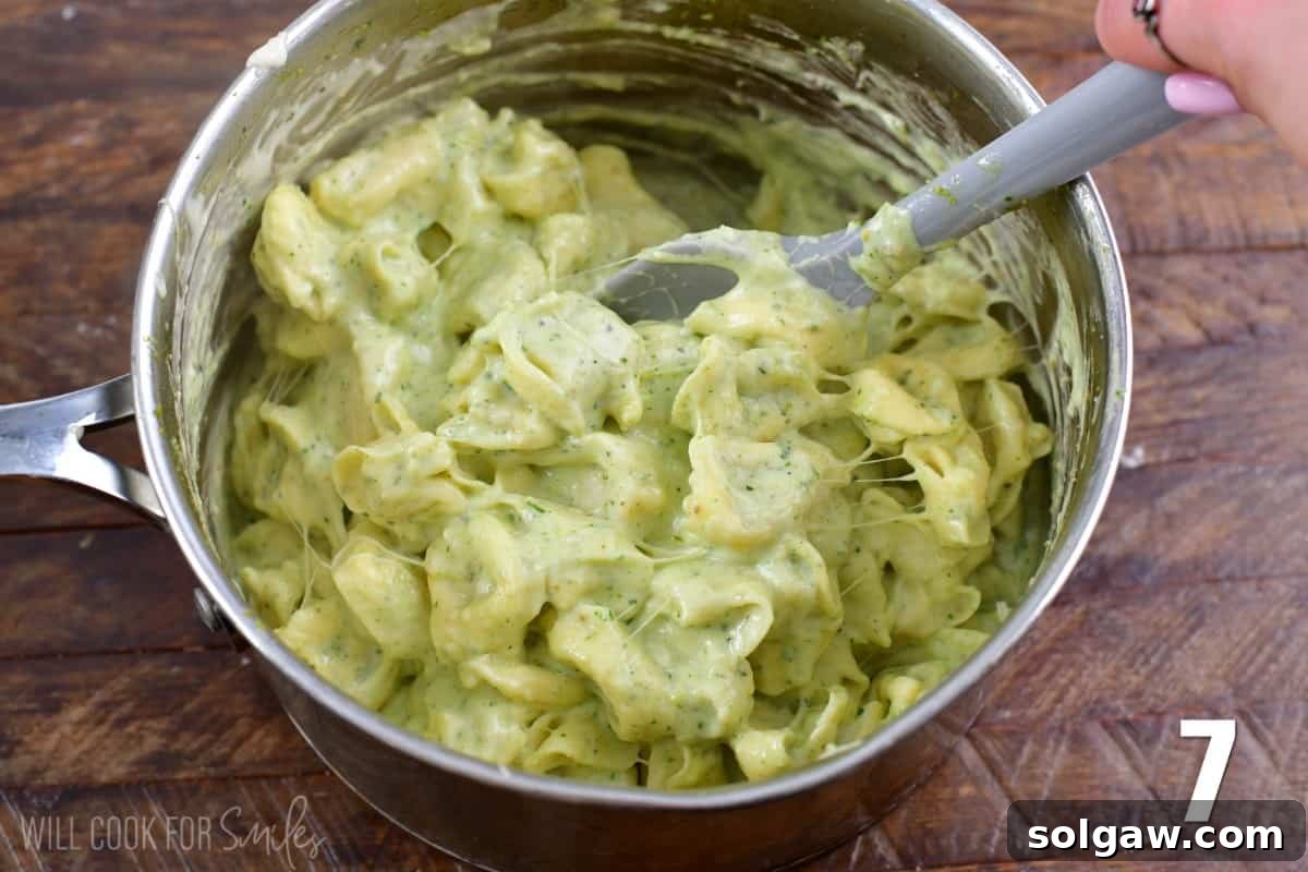 Creamy Pesto Alfredo Tortellini 6 Mixing pesto alfredo in a pan with the tortellini using a plastic spoon, showing the creamy, green coating.