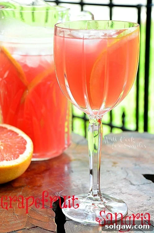 Grapefruit Sangria served in a wine glass, with half a grapefruit and a pitcher in the background, suggesting a refreshing drink.