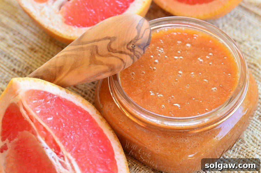 Glass jar filled with homemade grapefruit syrup and sauce on a tan placemat with a wooden spoon leaning on the jar and sliced grapefruit laying around the jar as viewed from above.
