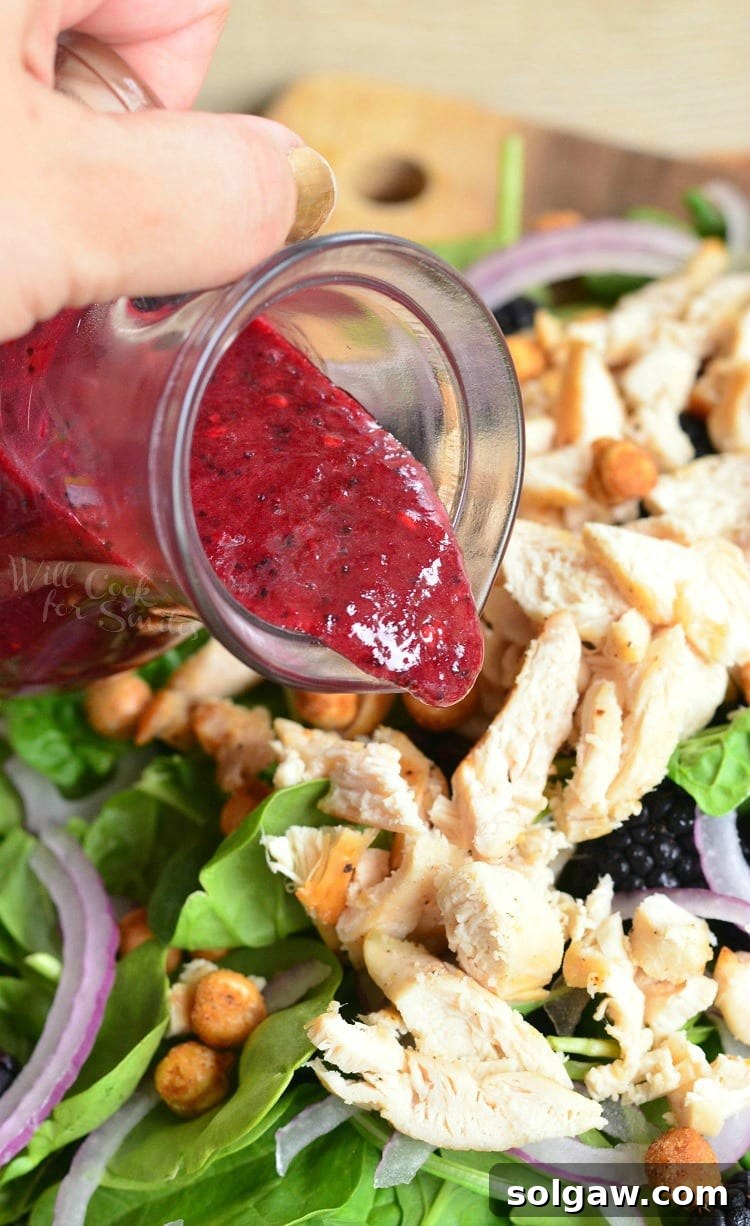 Blackberry Poppy Seed Chicken Spinach Salad 6 Pouring homemade Blackberry Poppy Seed Vinaigrette over a generous serving of Blackberry Chicken Spinach Salad, showcasing fresh lettuce, chicken, red onion, and blackberries.