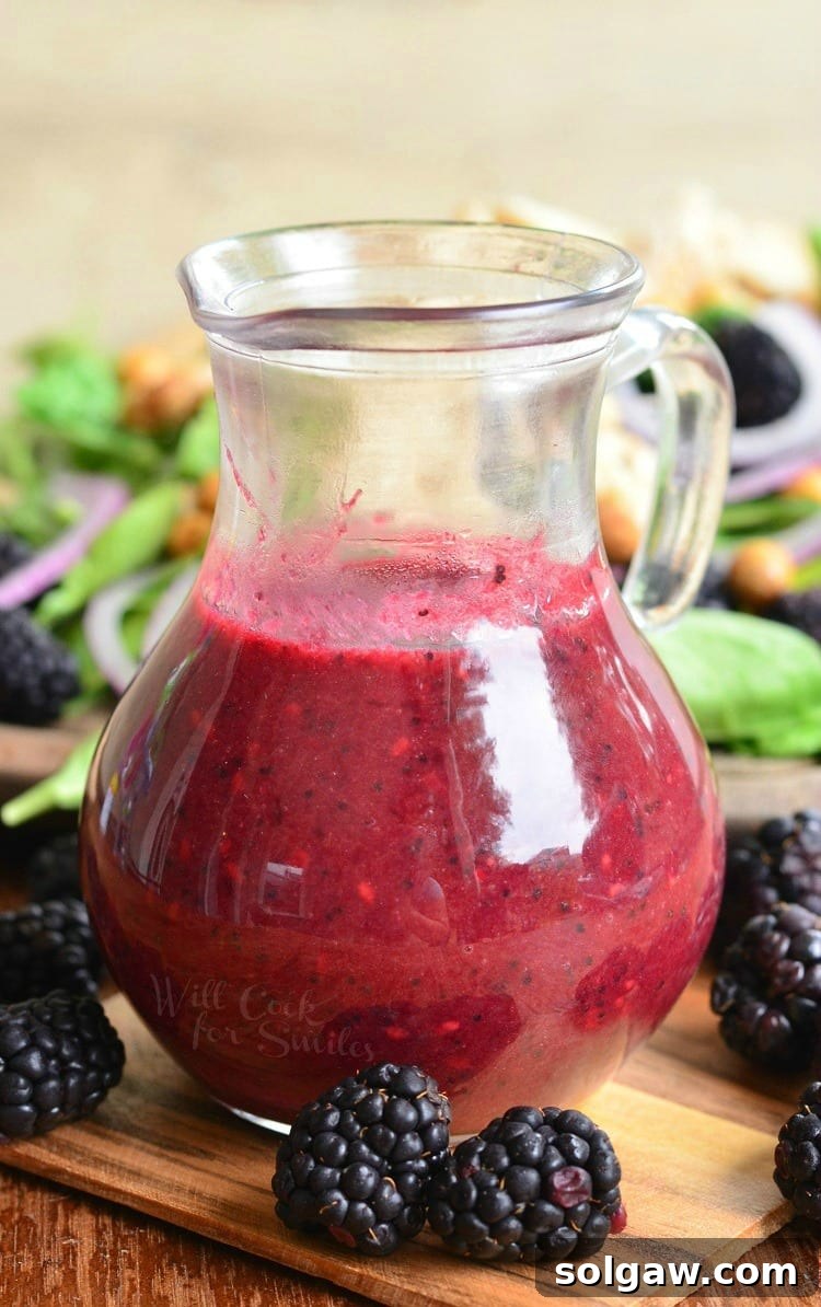 Blackberry Poppy Seed Chicken Spinach Salad 9 Blackberry Poppy Seed Vinaigrette in a glass container with blackberries around it on a cutting board, highlighting its rich color and texture.