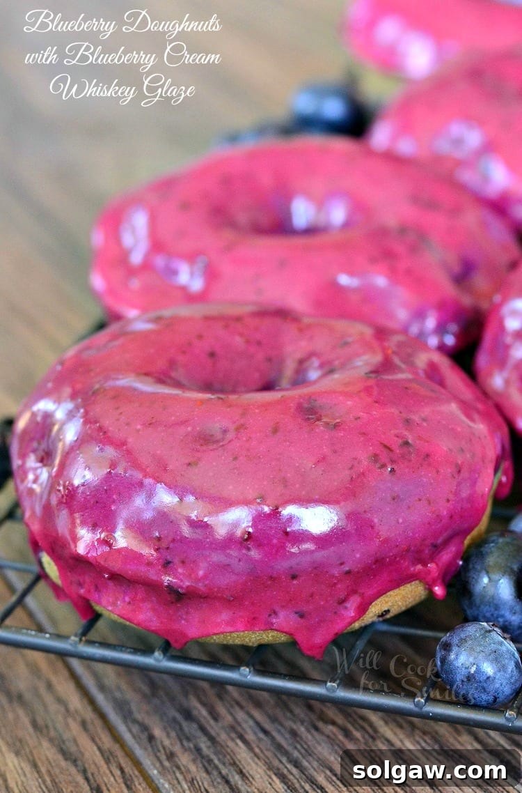 Close-up of baked blueberry doughnuts with luscious blueberry cream whiskey glaze on a cooling rack, fresh blueberries scattered around on a rustic wood table.