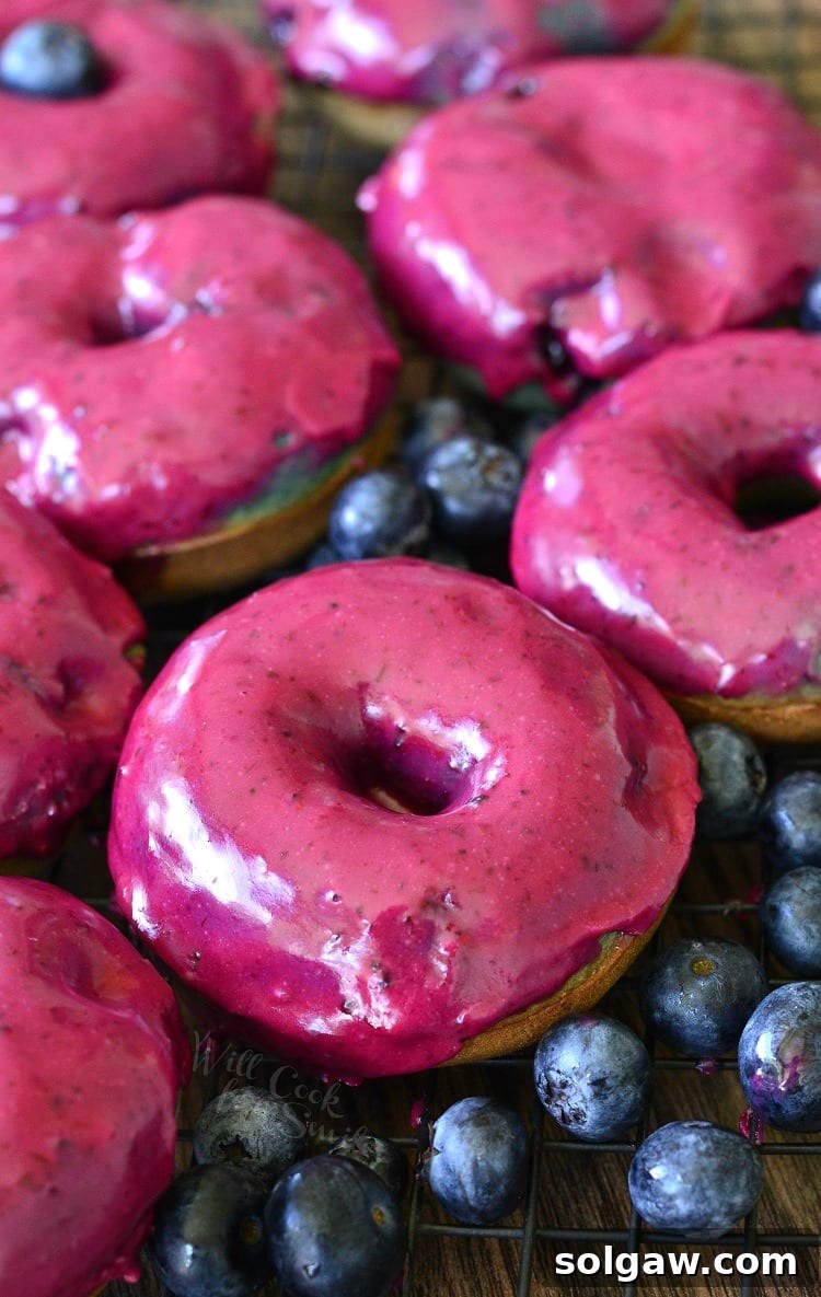 Baked blueberry doughnuts with blueberry cream whiskey glaze arranged on a wire cooling rack, surrounded by fresh blueberries on a wooden surface.