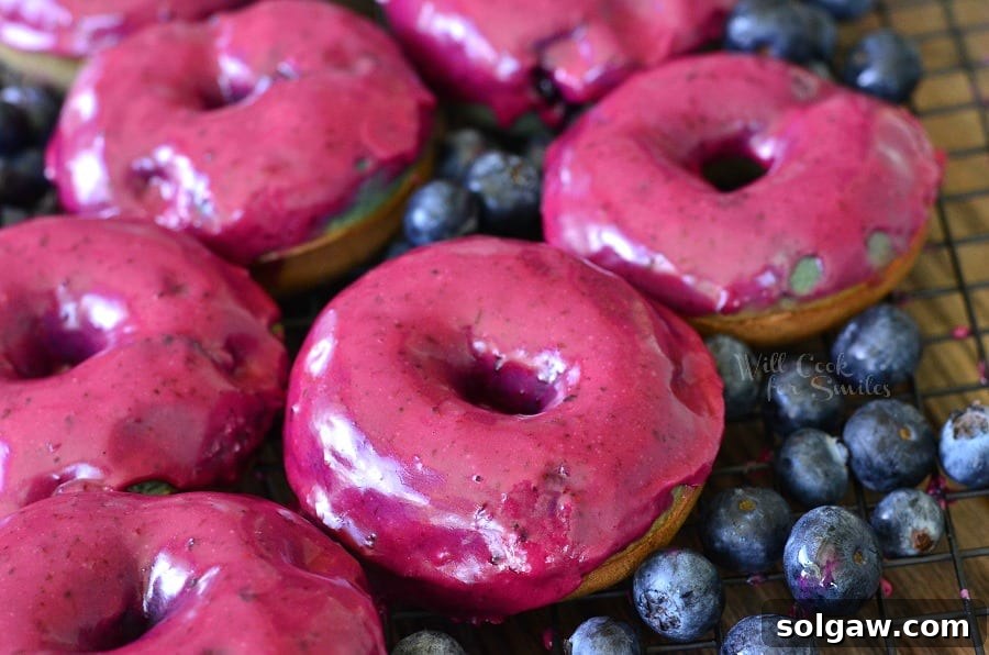 Artfully arranged baked blueberry doughnuts with thick blueberry cream whiskey glaze on a cooling rack, with scattered fresh blueberries, on a light wood table.
