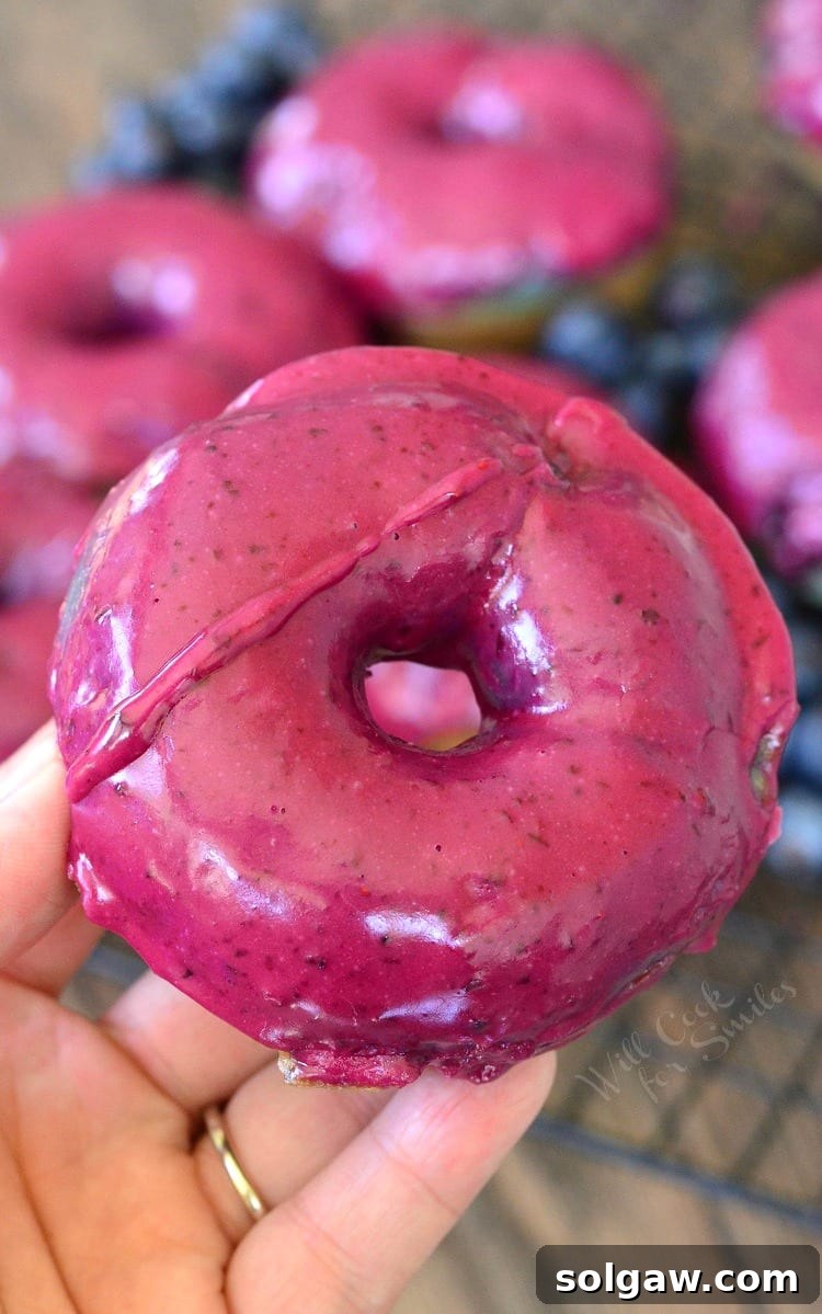 A person's hand holding a single, perfectly glazed baked blueberry doughnut, showcasing its rich topping and fluffy texture.