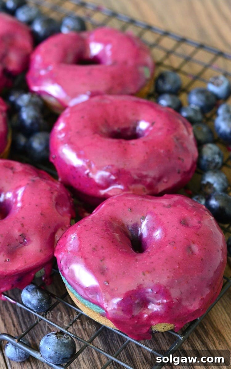 An overhead view of numerous baked blueberry doughnuts with a thick, glossy blueberry cream whiskey glaze, resting on a wire cooling rack on a wooden tabletop.