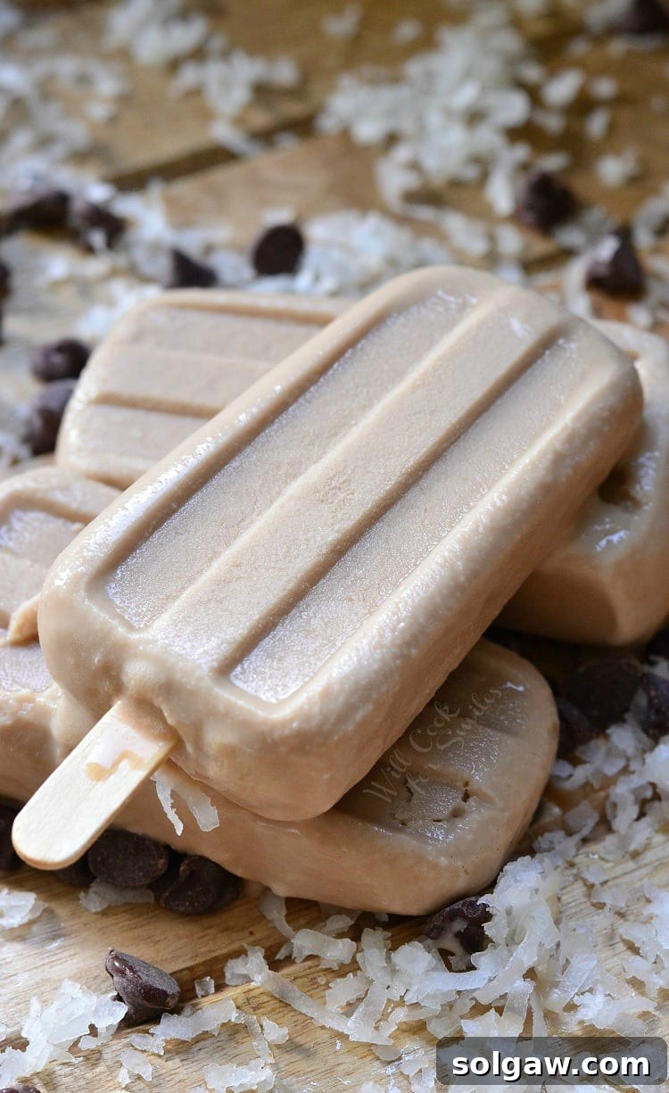 3 creamy coconut mocha ice pops stacked on each other while sitting on a wooden slatted table with coconut and chocolate chips scattered around the table