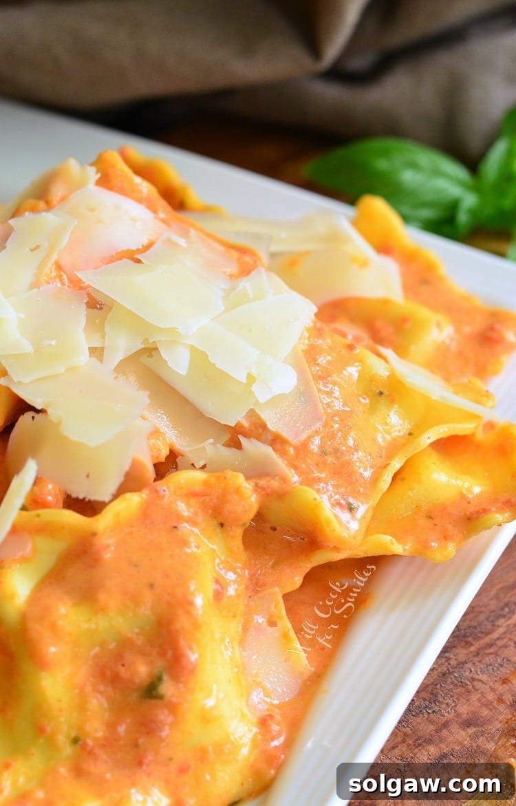 Decorative white square plate filled with ravioli in creamy red pepper sauce topped with shaved parmesan.