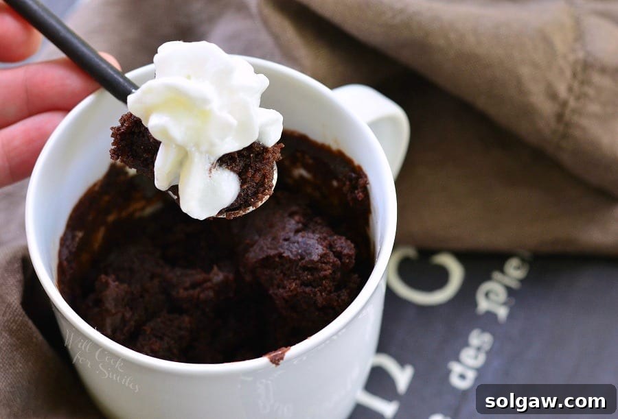 Chocolate cake in a white coffee mug with a spoon lifting some out with whip cream on top 