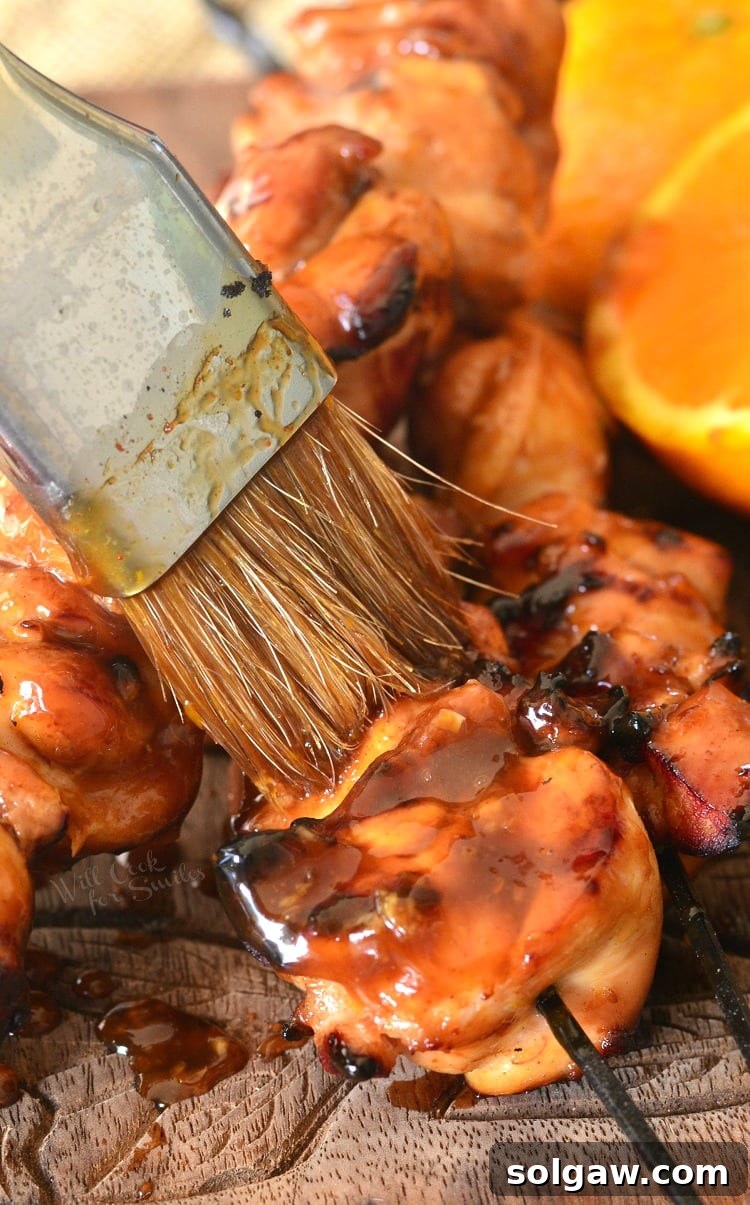 Orange Glazed Chicken Skewers 8 Close-up of a hand brushing sticky orange sauce onto cooked chicken thigh kebabs on a wooden cutting board.