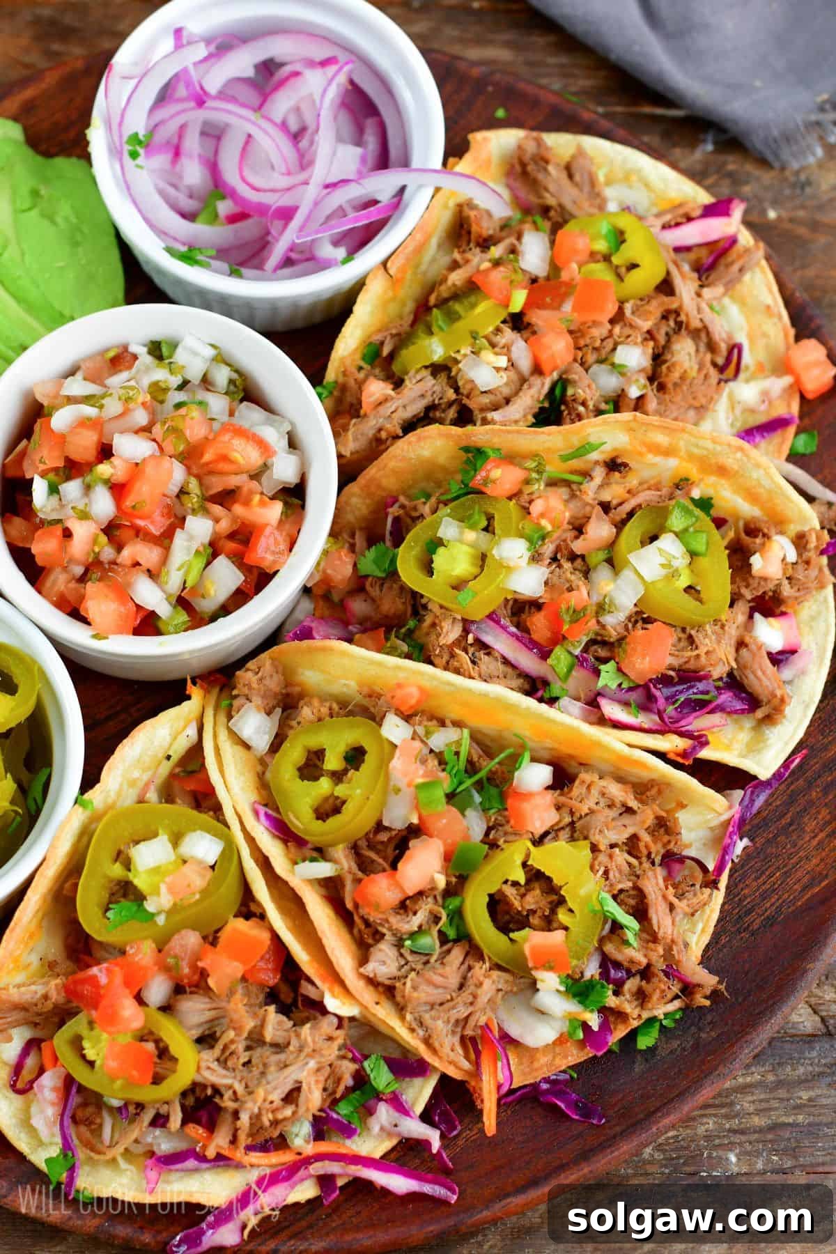 Four pulled pork tacos beautifully arranged on a rustic wooden plate, accompanied by small bowls of vibrant red onion, fresh Pico de Gallo, and tangy pickled jalapenos, ready to be enjoyed.