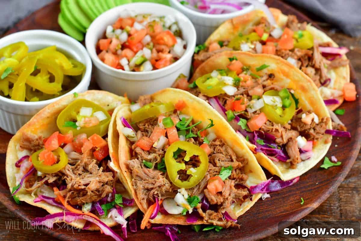 Four beautifully assembled pulled pork tacos artfully arranged on a wooden plate, surrounded by small bowls brimming with fresh tomato, vibrant Pico de Gallo, and zesty pickled jalapenos as toppings.