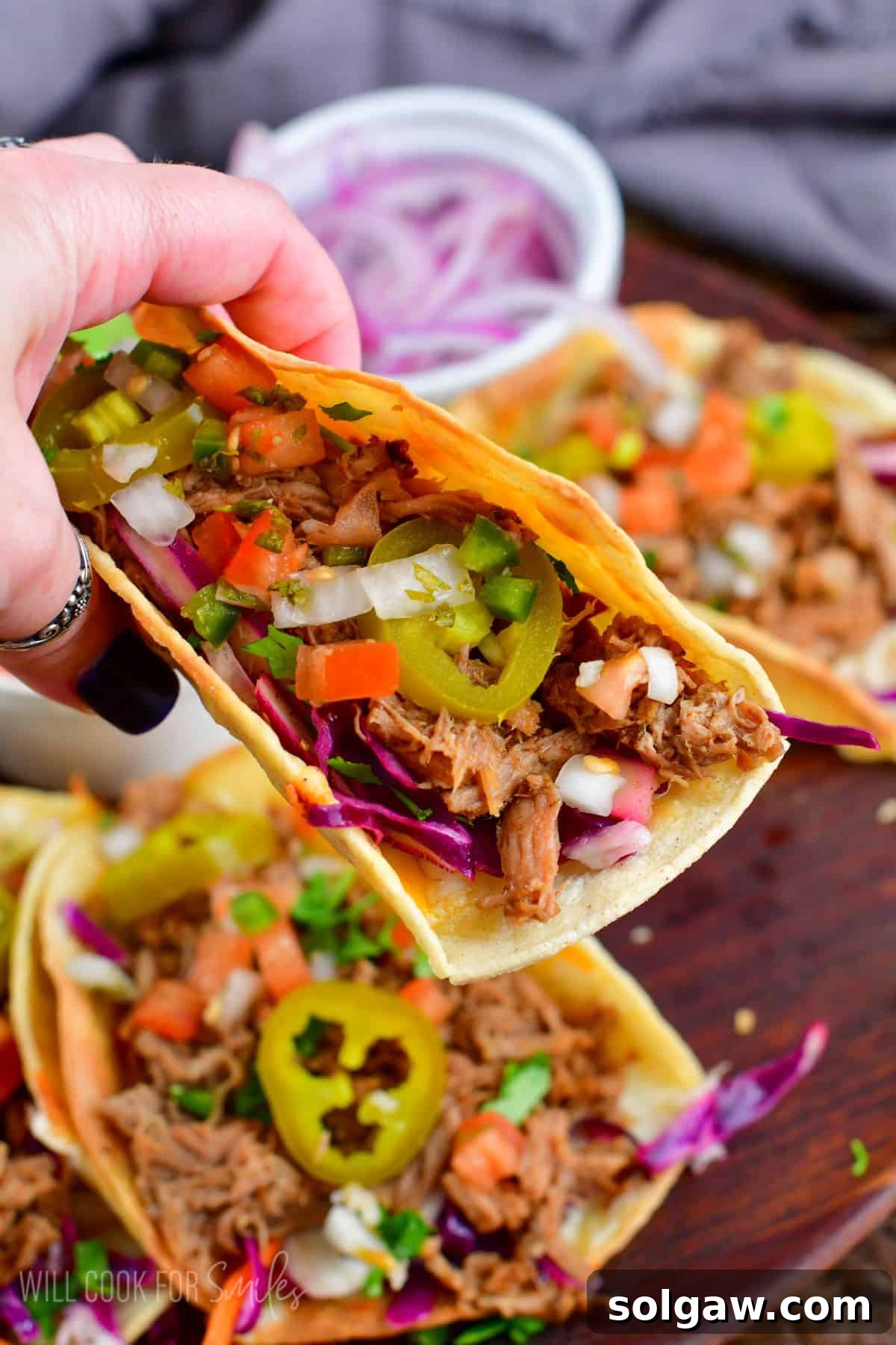 A close-up shot of a hand holding a perfectly assembled pulled pork taco, with more tacos visible in the background, showcasing the delicious layers of meat and toppings.