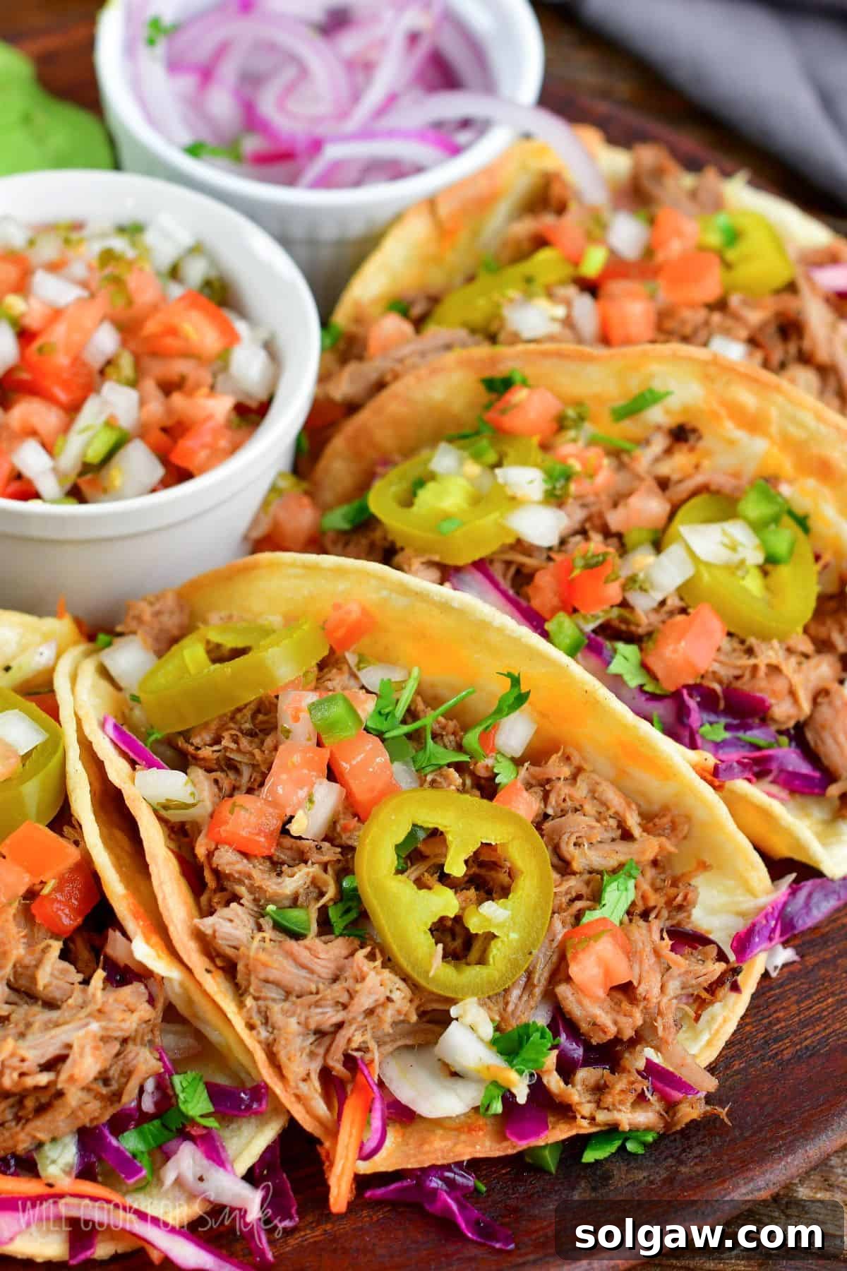 Four delicious pulled pork tacos arranged on a wooden plate, artfully topped with pickled jalapenos, fresh diced tomato, and vibrant Pico de Gallo for a burst of flavor.