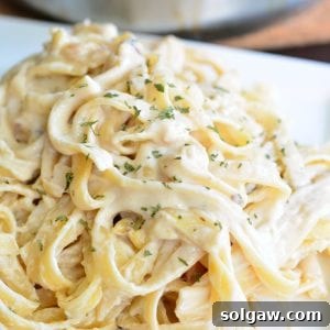 A top-down shot of Roasted Garlic Asiago Fettuccine in a metal bowl, garnished with fresh parsley and pine nuts, with metal tongs ready to serve.