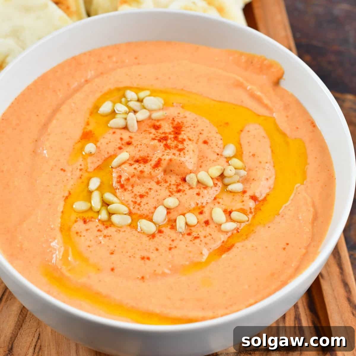 Squared off image of the creamy pinkish-red roasted red pepper hummus in a white bowl, garnished with olive oil and a few red pepper flakes.