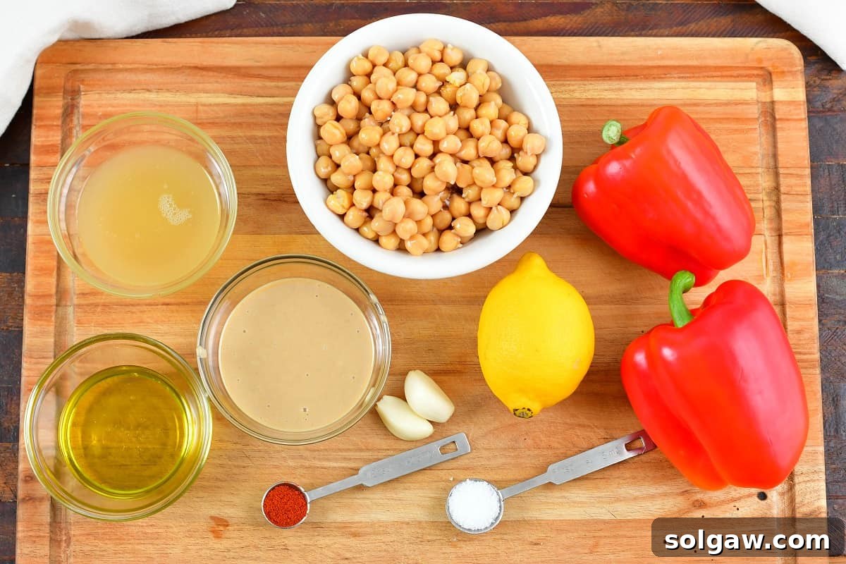 Assorted ingredients for roasted red pepper hummus, including chickpeas, olive oil, tahini, lemon, garlic, and red bell peppers, arranged on a wooden board.