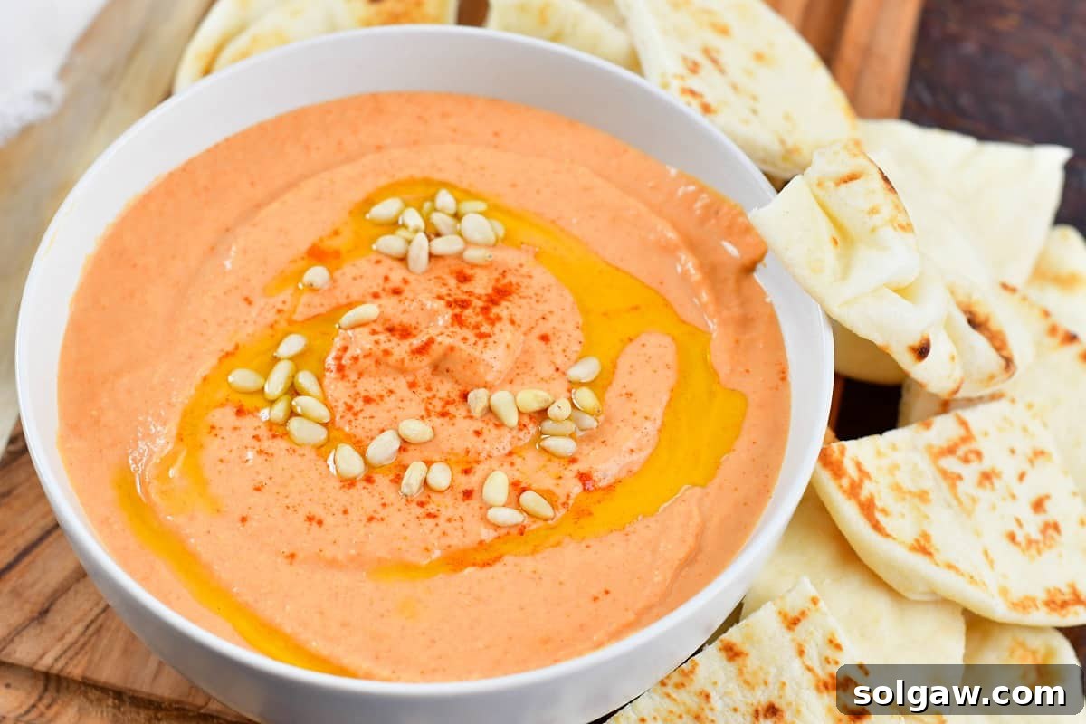 Close-up of vibrant pinkish-red roasted red pepper hummus in a white bowl, garnished with a swirl of olive oil.