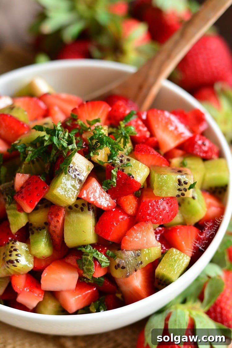 A crisp white bowl filled with a delightful mix of chopped strawberries and kiwi, garnished with fresh mint leaves. A wooden spoon rests inside, ready to serve.
