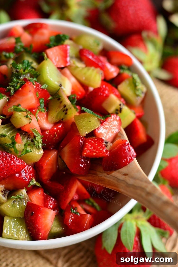 Close-up shot of a strawberry and kiwi fruit salad in a white bowl, with a wooden spoon scooping out a serving, showcasing the vibrant colors and fresh mint garnish.