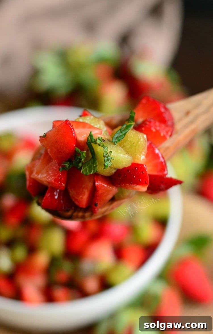 A close-up of a wooden spoon lifting a spoonful of strawberry and kiwi fruit salad from a pristine white bowl, highlighting the fresh mint.