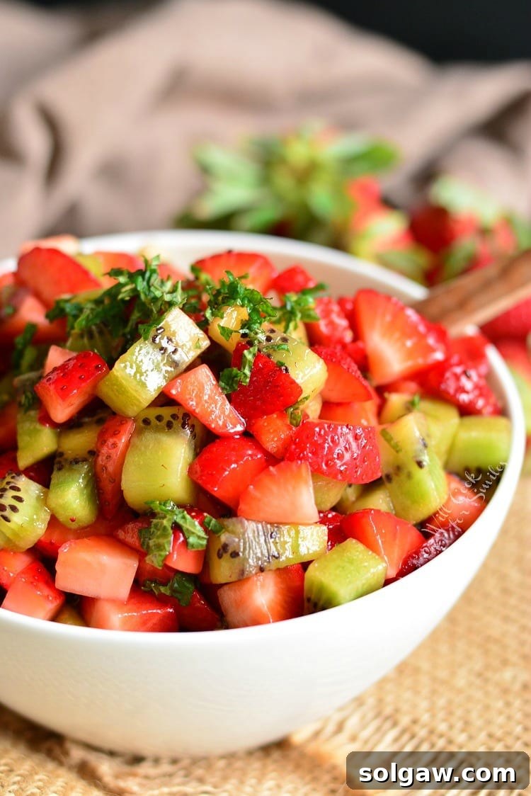 A beautiful close-up of the strawberry and kiwi fruit salad, garnished with vibrant green mint leaves, in a white bowl.