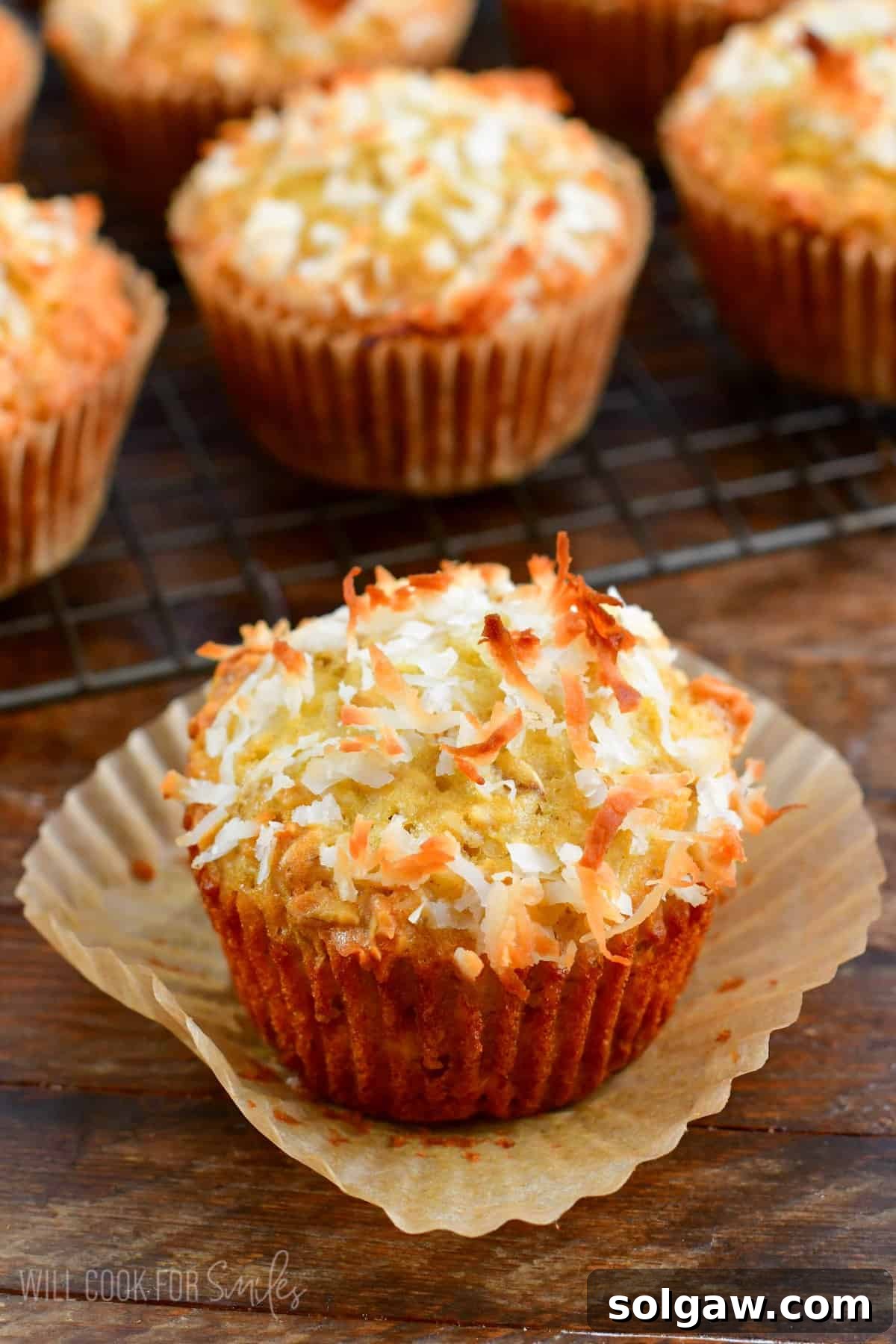 A freshly baked Coconut Oat Muffin with its liner removed, revealing a soft, textured interior. More muffins are cooling on a wire rack in the background.