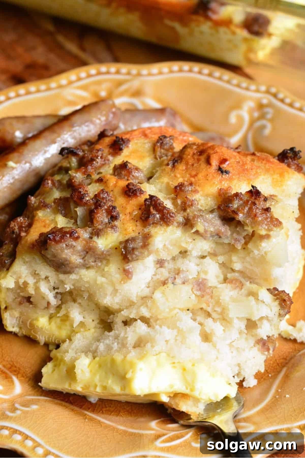 Hearty Sausage & Egg Breakfast Bake 8 A close-up shot of a slice of sausage breakfast casserole on a plate with additional sausage crumbles and a fork taking a bite.