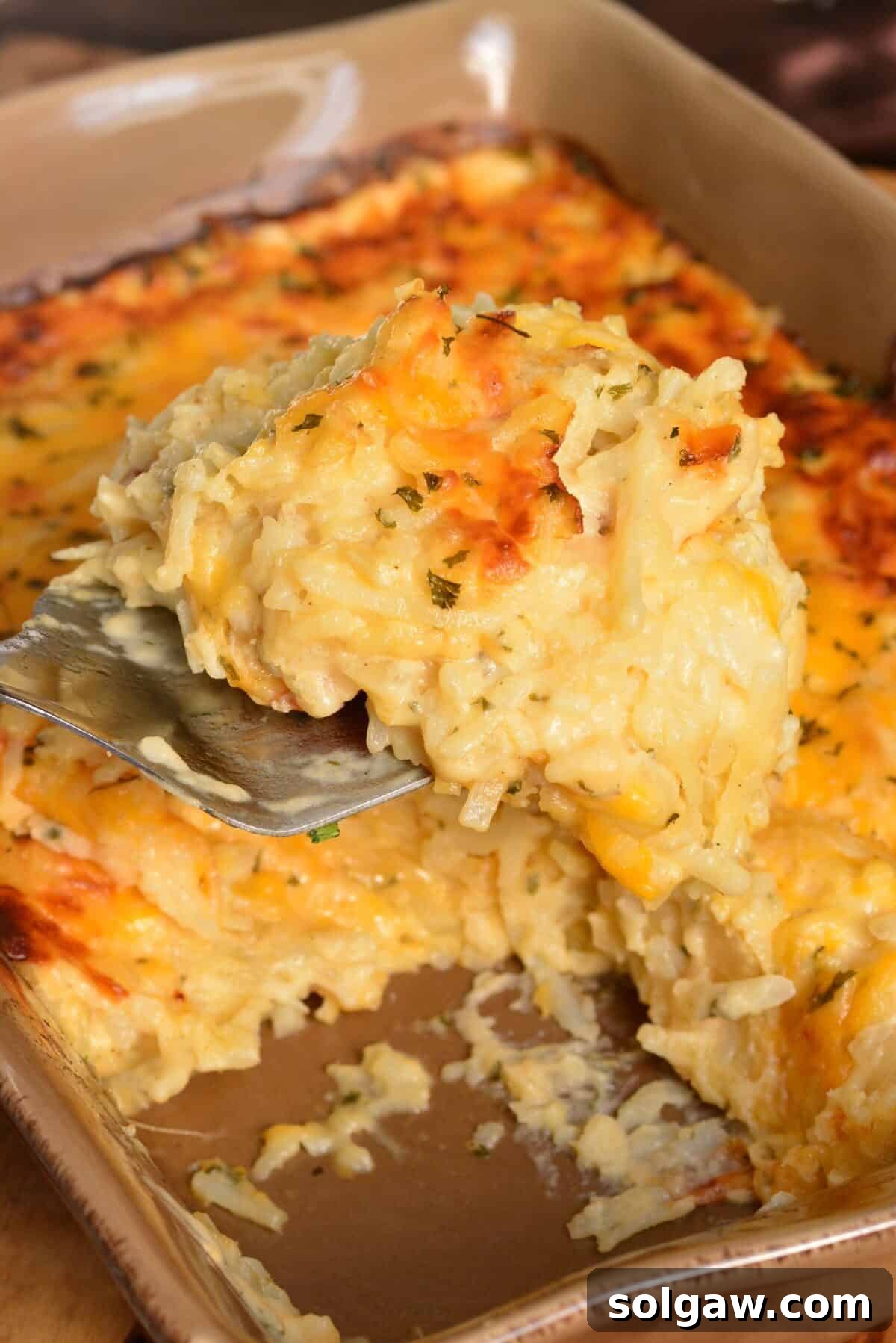 Scooped portion of cheesy hashbrown casserole on a metal spatula over the baking dish, highlighting its gooey texture.