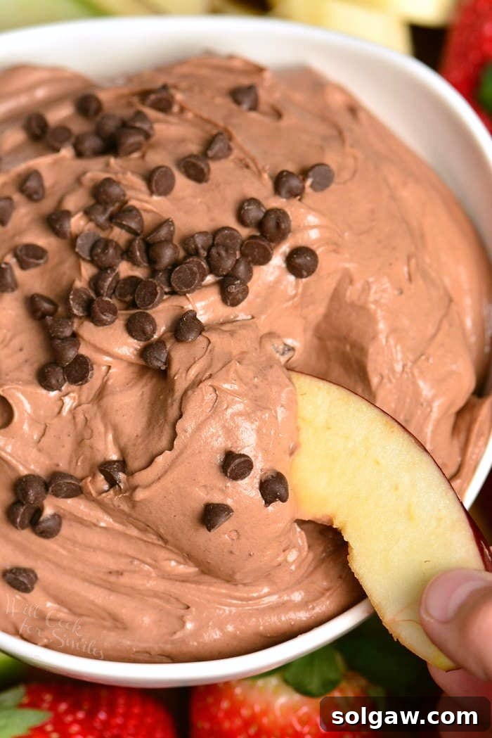 chocolate cake dip in a white bowl with chocolate chips on top and a apple slice being dipped in the bowl.