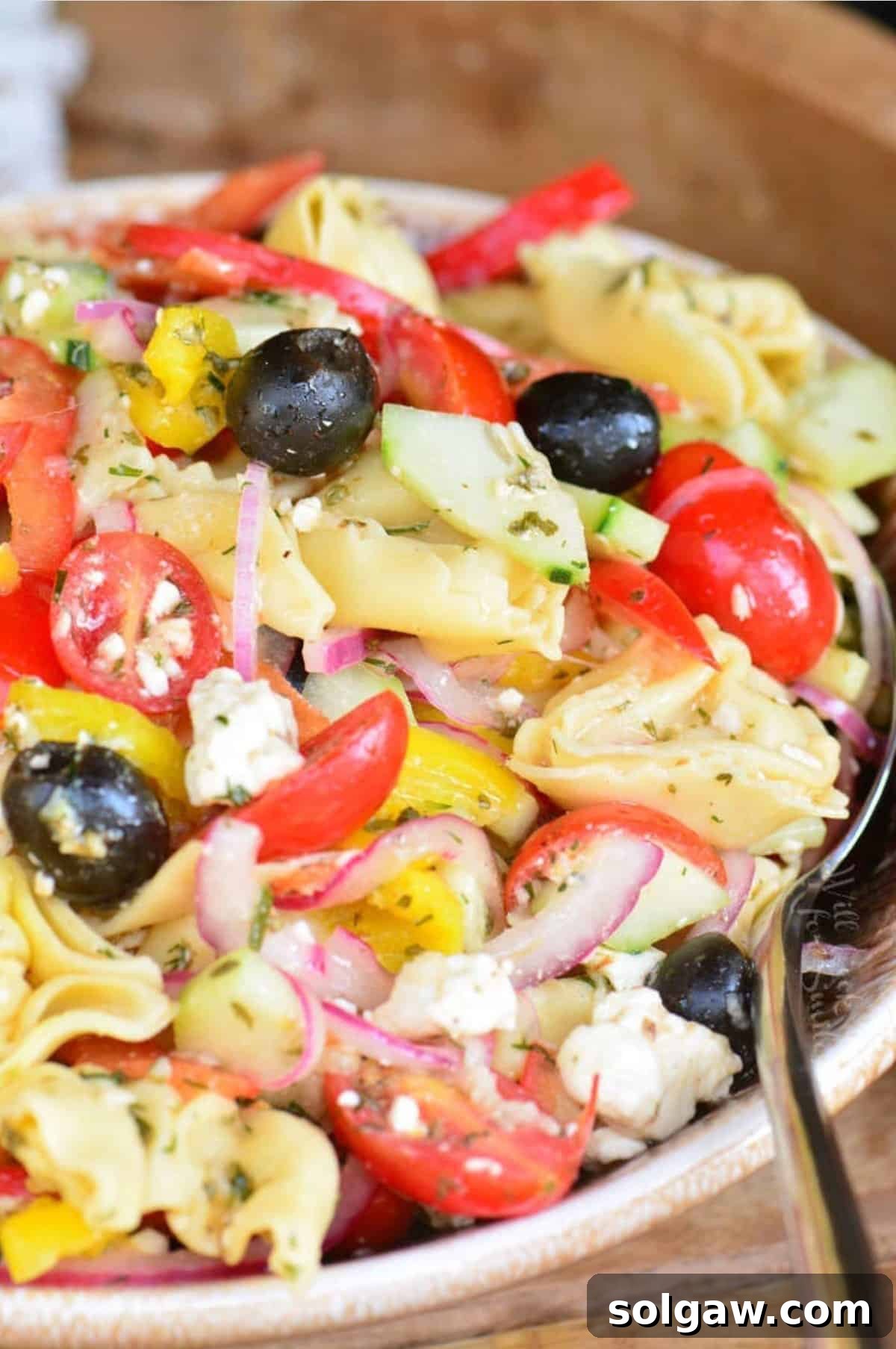 A bowl of vibrant Greek Tortellini Salad with a large serving spoon, showcasing fresh vegetables, creamy feta, and tender tortellini.