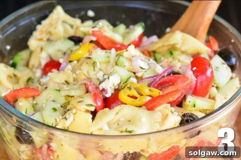 A close-up shot of a large glass bowl filled with Greek Tortellini Salad, being gently mixed with a wooden spoon.