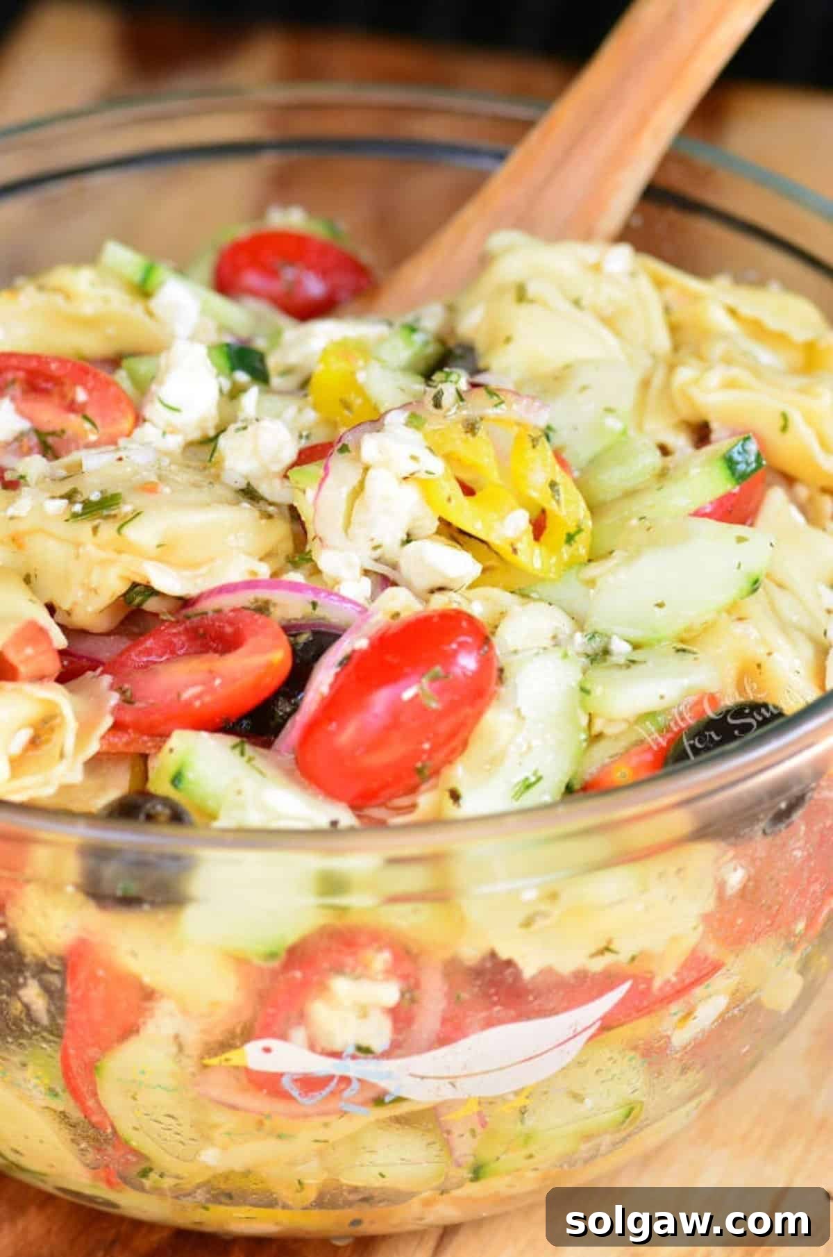 A bountiful glass bowl of mixed Greek Tortellini Salad, ready to serve, accompanied by a wooden serving spoon.