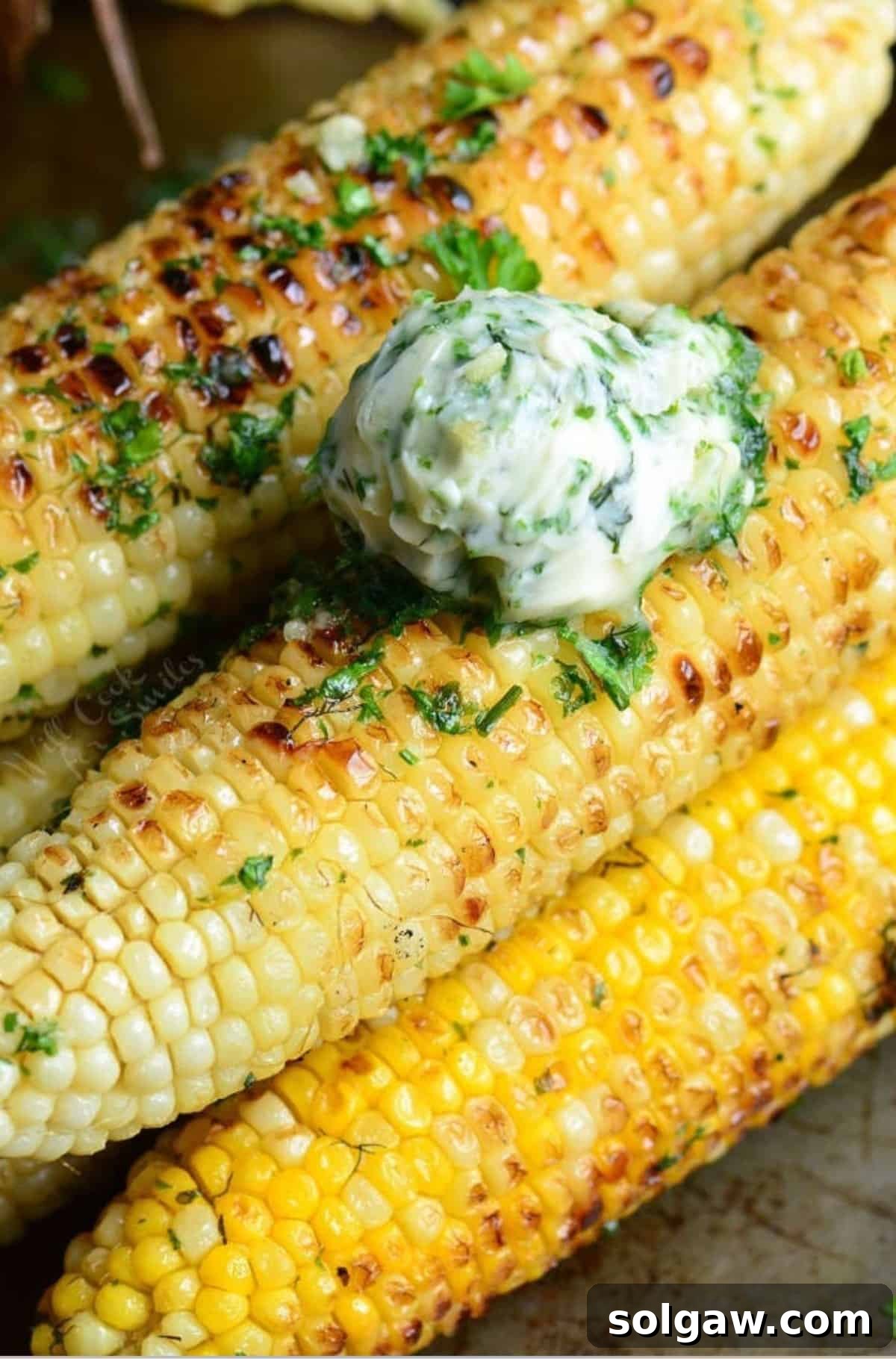 Several stacked grilled corn on the cob, generously topped with a scoop of homemade garlic herb butter, creating an inviting and flavorful presentation.
