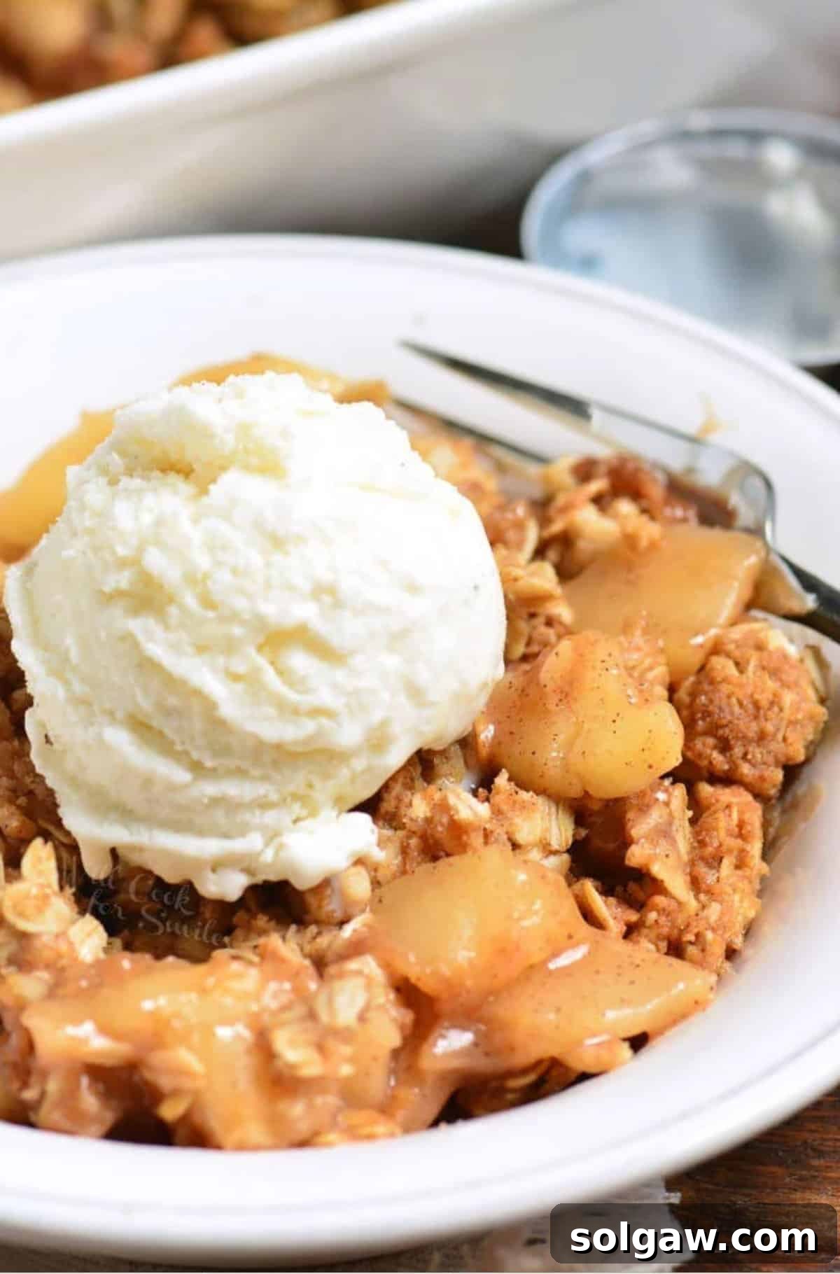 A white bowl of apple crisp topped with a large scoop of vanilla ice cream, ready to be enjoyed.
