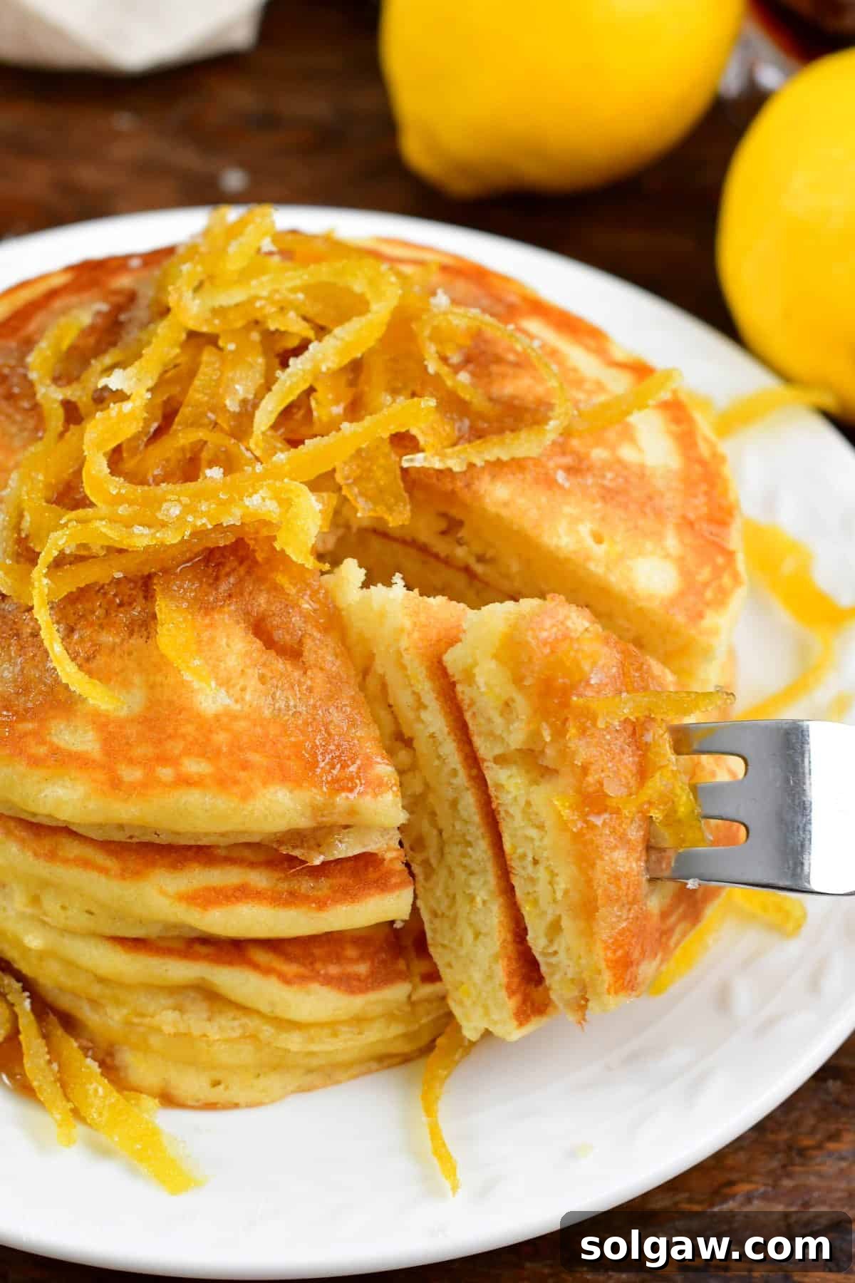 Bright Lemon Pancakes 2 Golden lemon pancakes stacked high with a slice cut out, being lifted with a fork, garnished with fresh lemon zest.