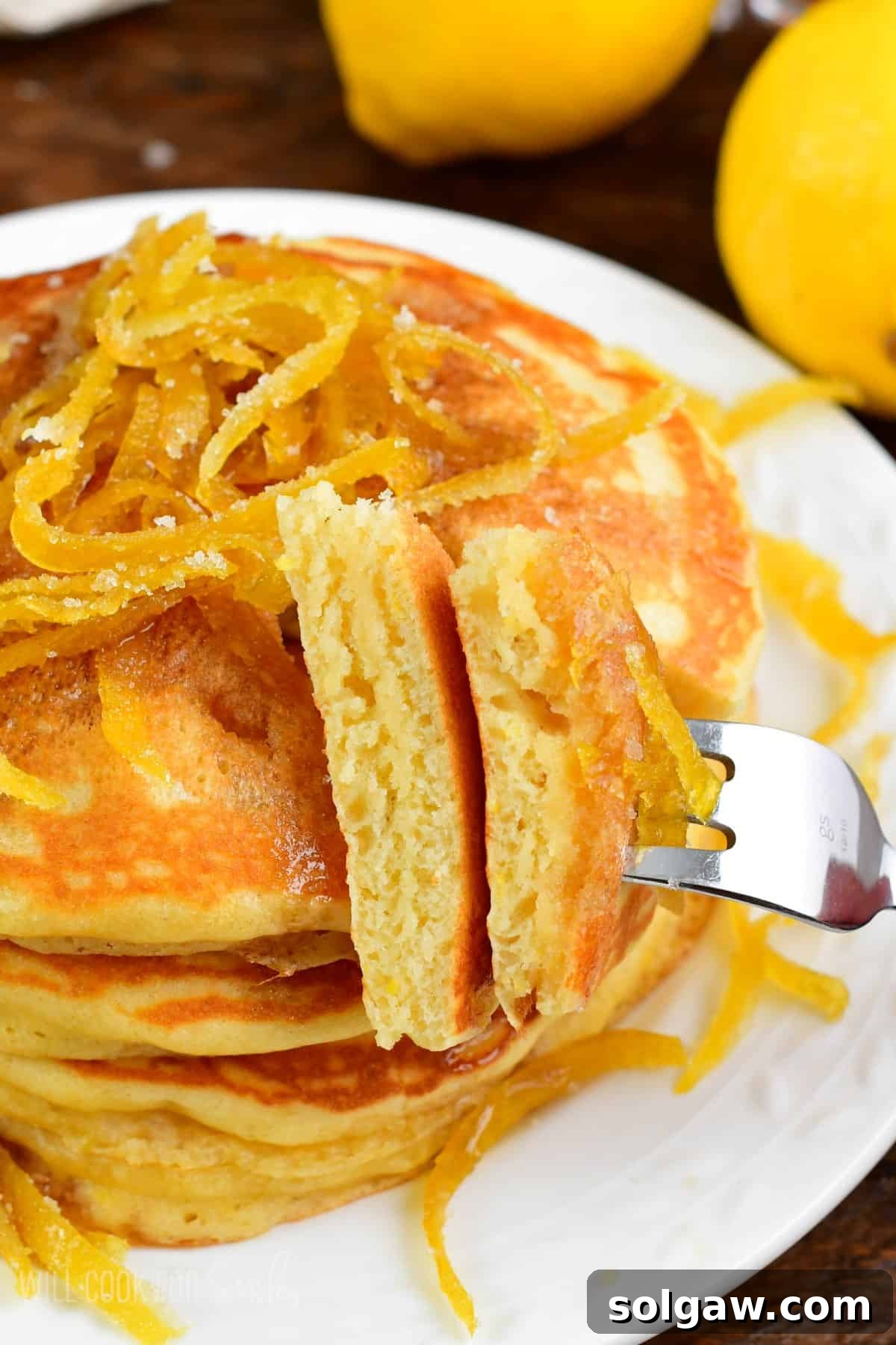 Bright Lemon Pancakes 6 Close-up of fluffy lemon pancakes with lemon peels on top, showing a piece lifted on a fork, highlighting their soft texture.