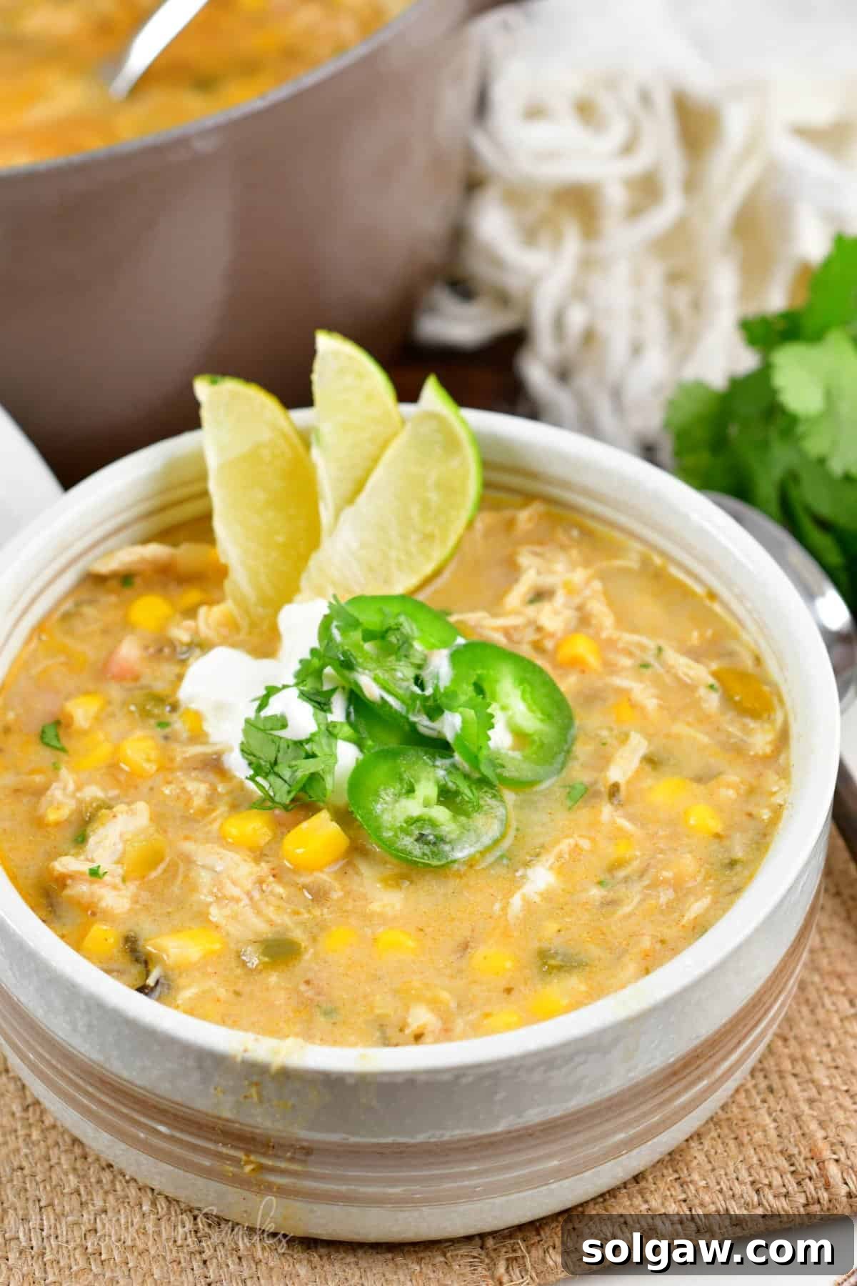 Creamy White Chicken Chili 2 white chicken chili in a bowl with three sliced limes, jalapeno, and sour cream on top on a burlap placemat.